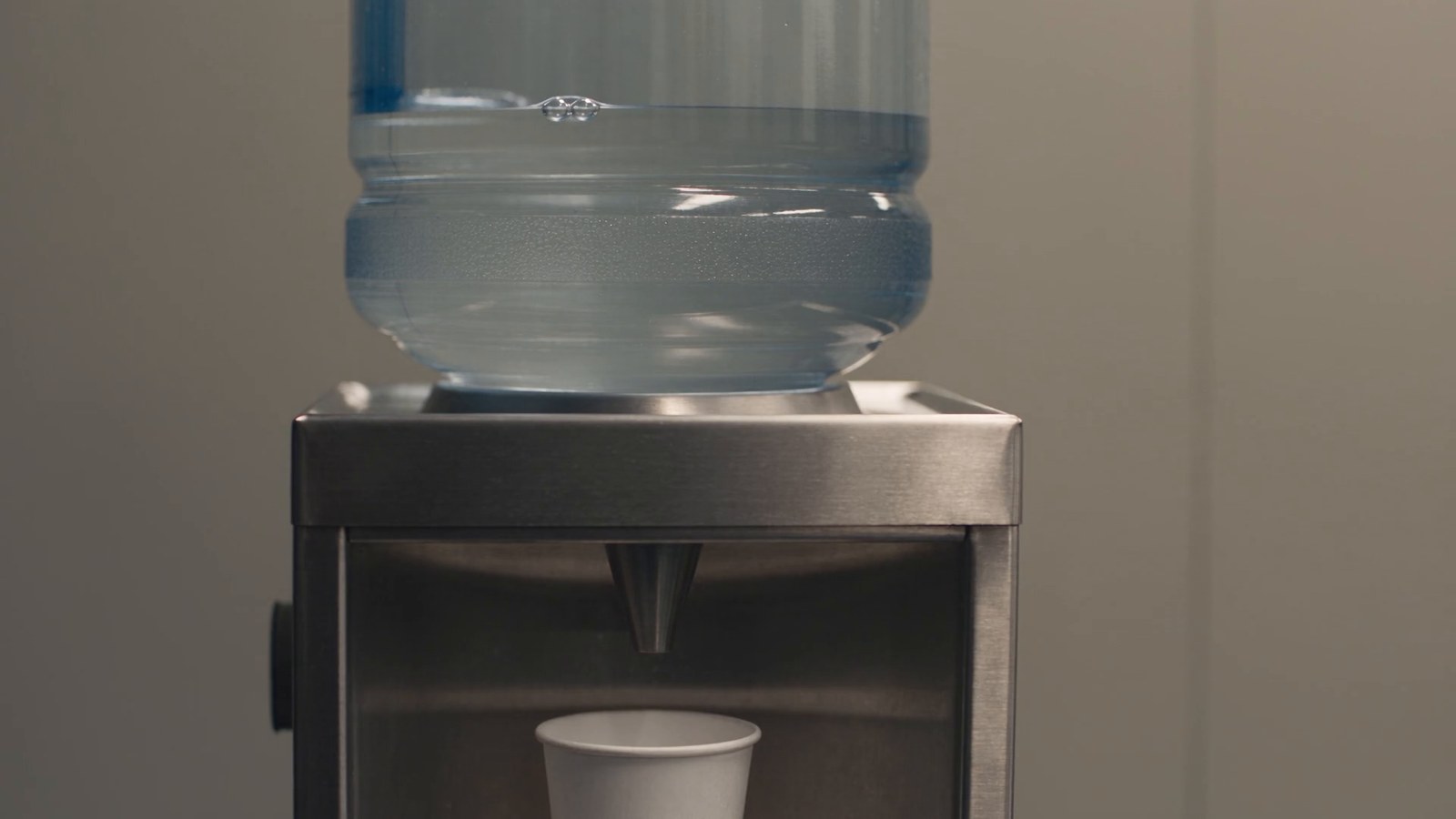 An office water cooler with its bottle nearly empty and a paper cup under the tap.