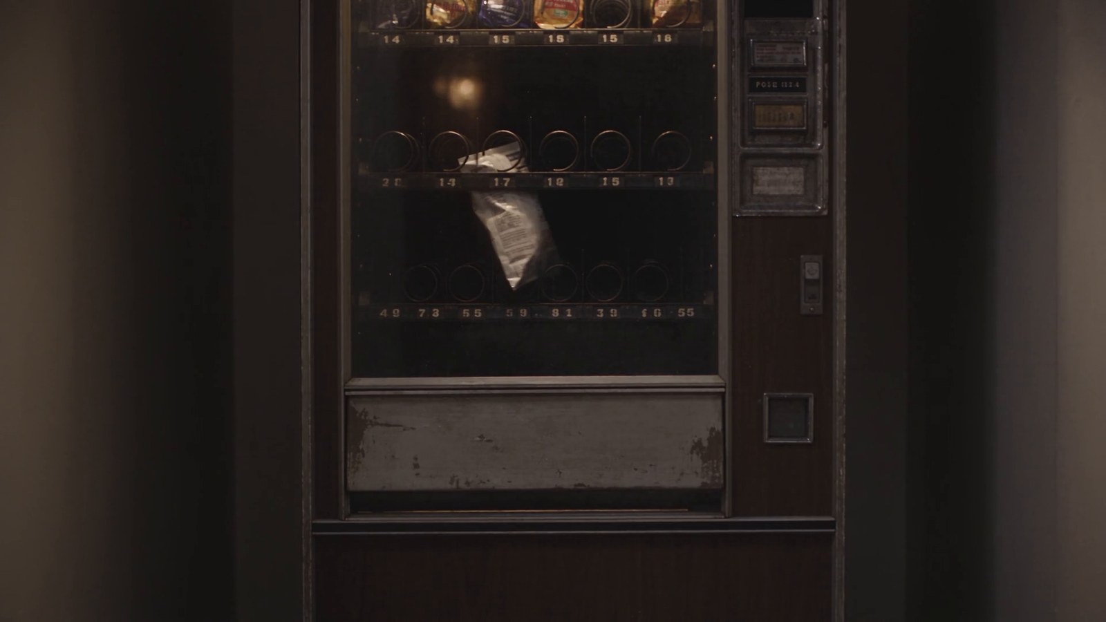 A snack bag hangs caught on the coil of an old vending machine in a dim hallway.